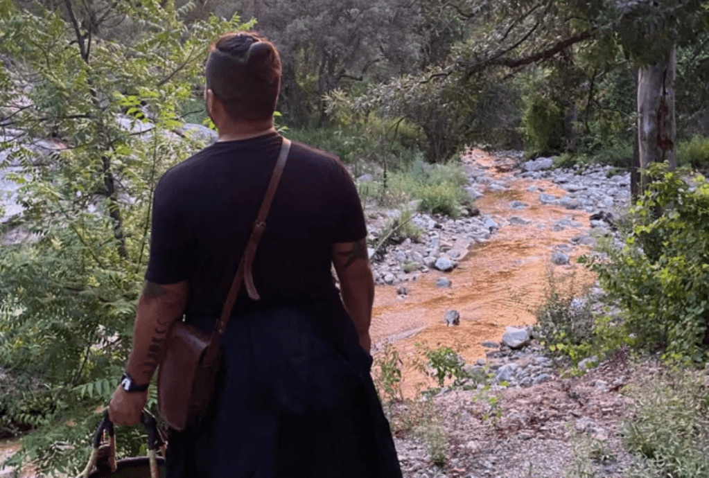 a man with a basket slung over a shoulder looks into the woods