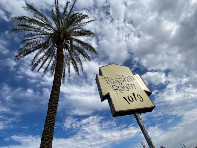 A sign for a music venue next to a palm tree underneath a blue sky with clouds.
