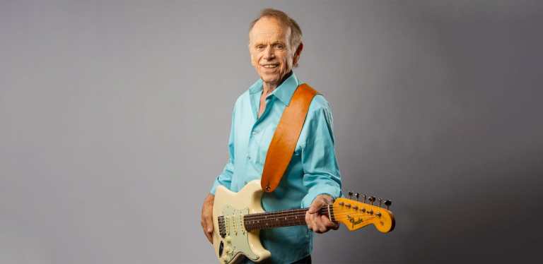 Older male in an aqua-colored short holding an electric guitar.