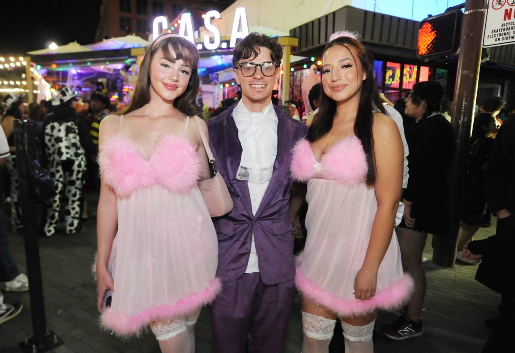 A man dressed as Austin Powers stands with two women dressed as Fembots on a crowded sidewalk.