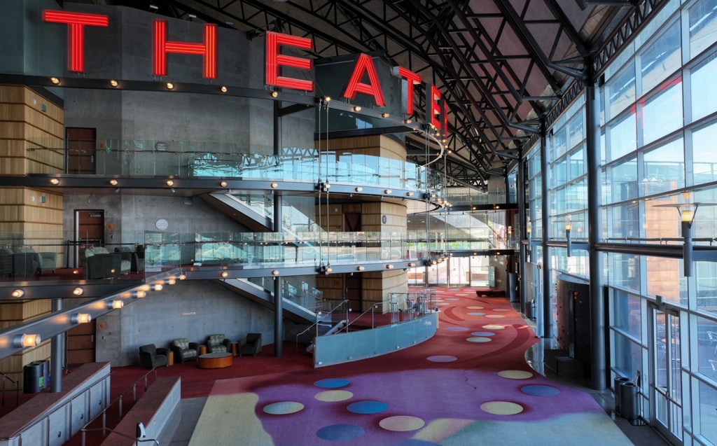 An interior view of the multi-story lobby of a performing arts center with large glass windows on one sides.