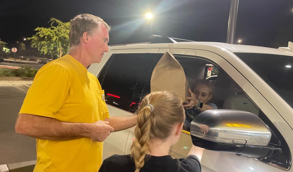 a man and young girl hand a food order to a woman in the passenger seat of a car