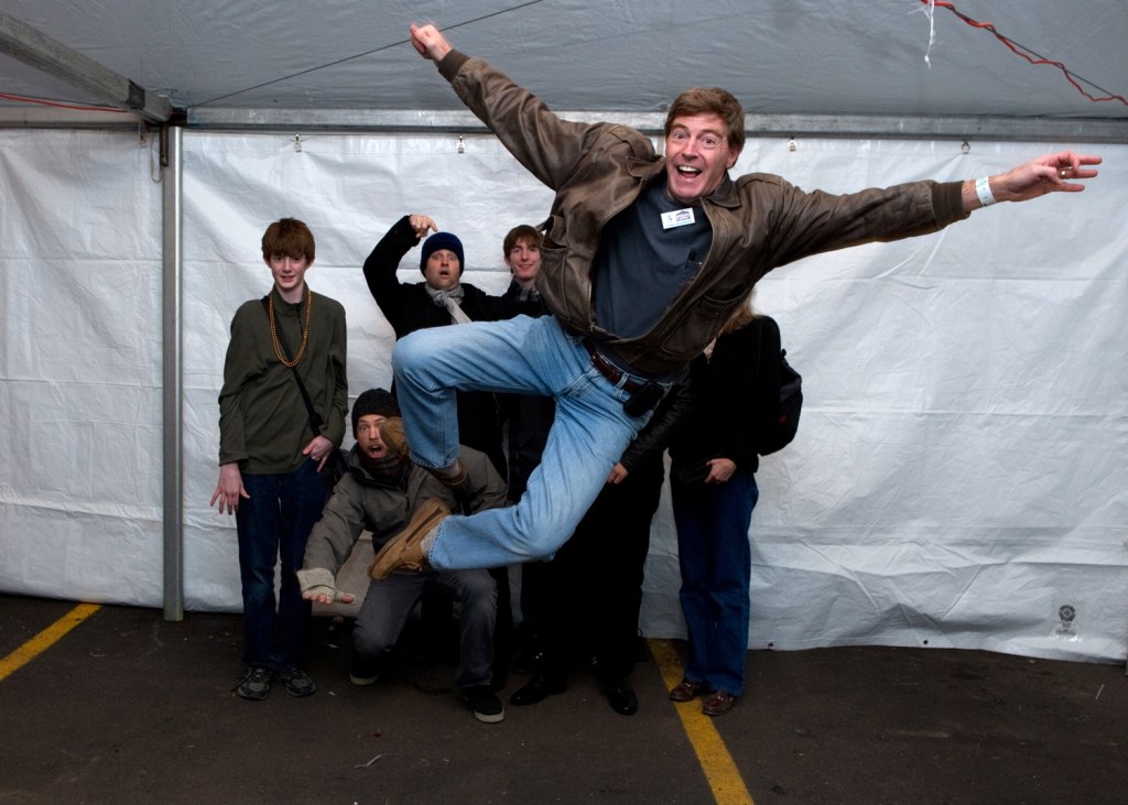 A middle-aged man jumps in the air in front of a group of people.