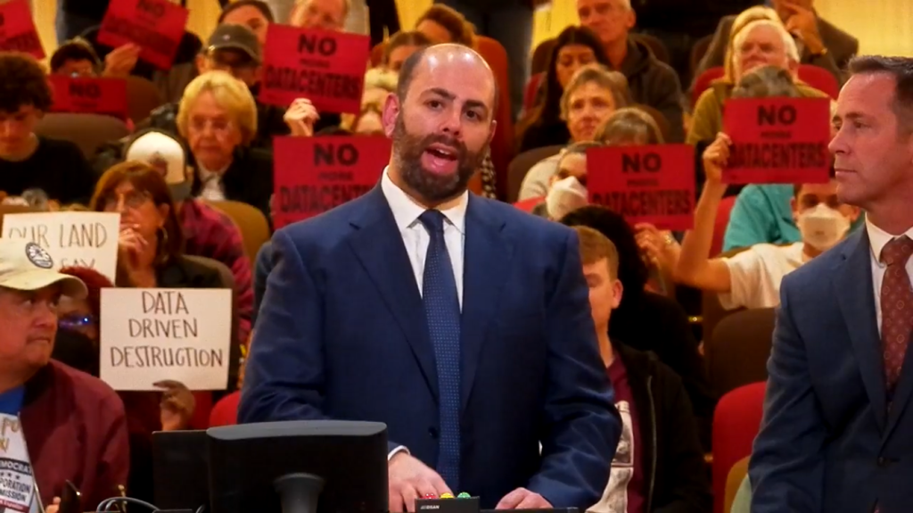 two men in suits speak in front of an audience at a city council meeting