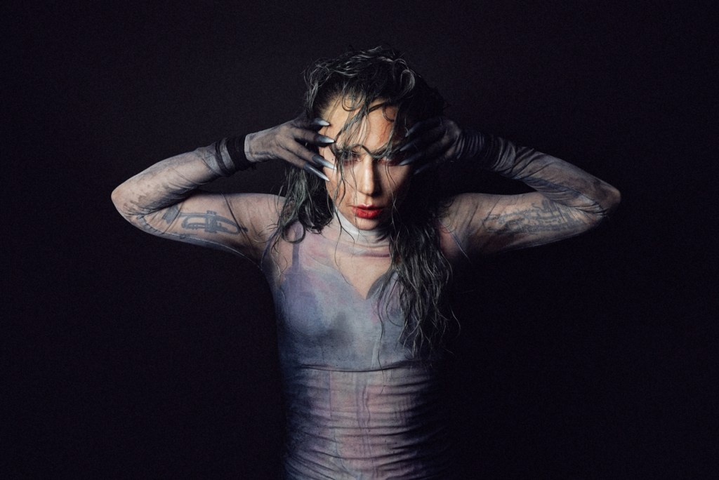 A long-haired woman in a mesh top and bra looks directly at the camera.