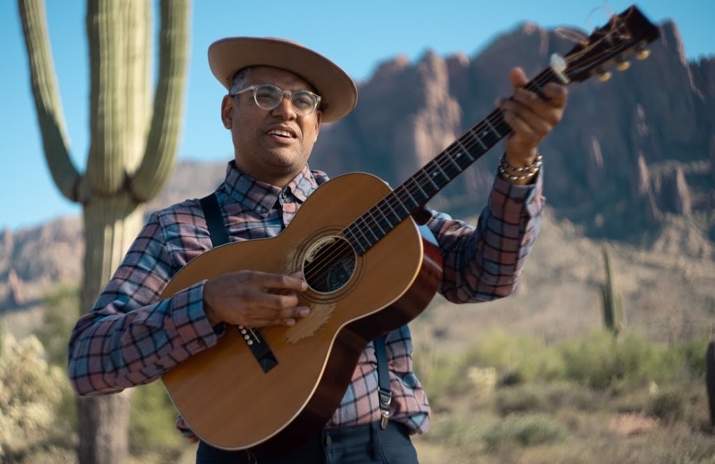 A man in glasses, a checkered shirt and a hat holds a guitar in the desert.