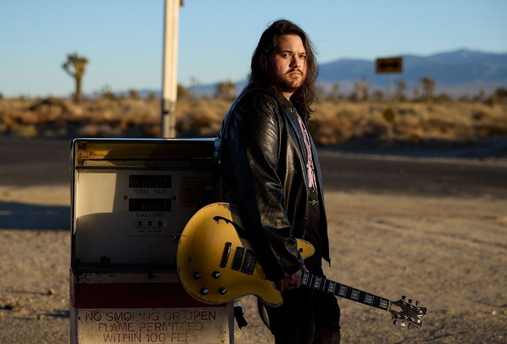 A long-haired musician holding a guitar at a desert gas station.