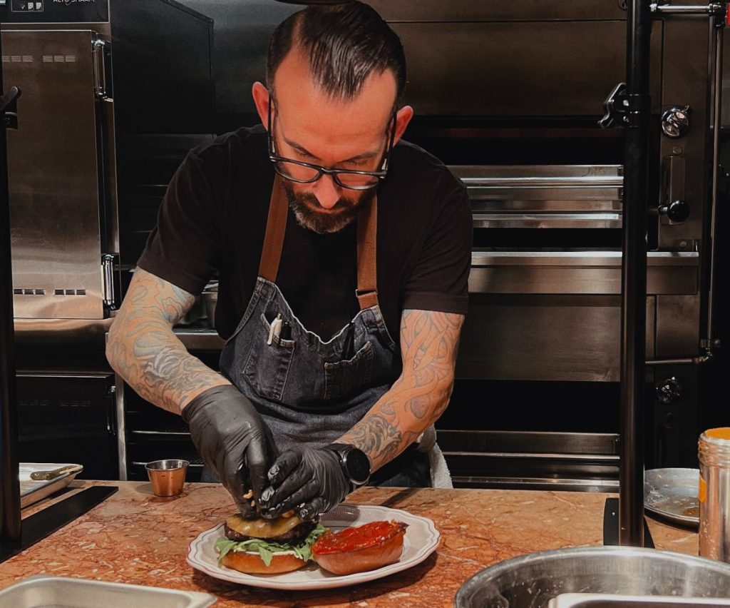 A chef adds toppings to a plated burger.