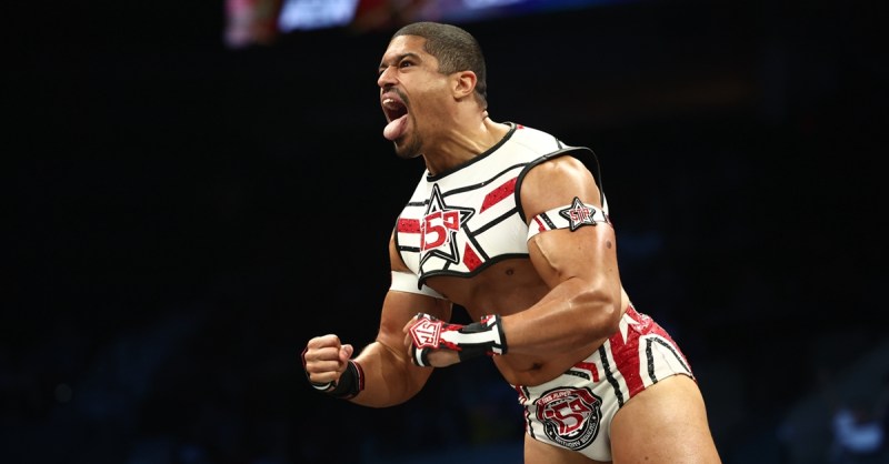 Anthony Bowens poses and shouts during his entrance before an AEW match, wearing a red-and-white crop top and matching ring gear inside the arena.