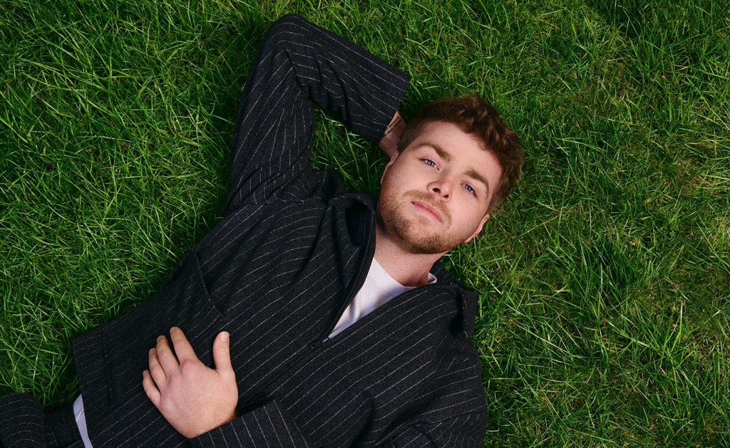 YouTuber and pop singer Alex Warren lays in the grass while looking at the camera.