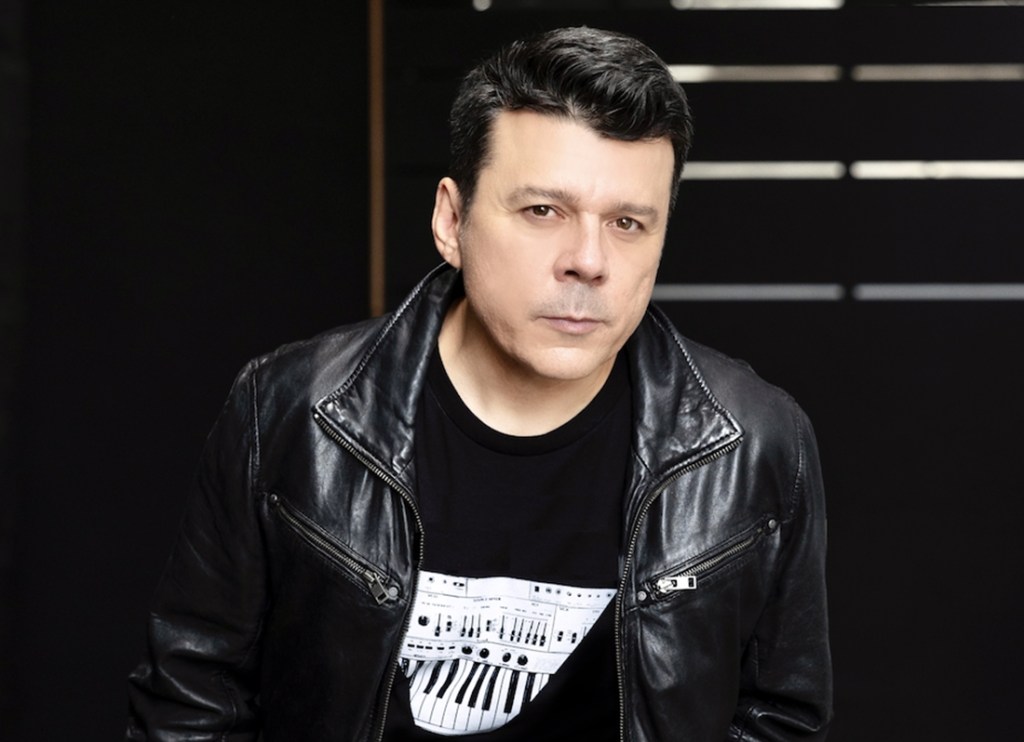 A black-haired man in a black leather jacket and t-shirt looks directly at the camera.