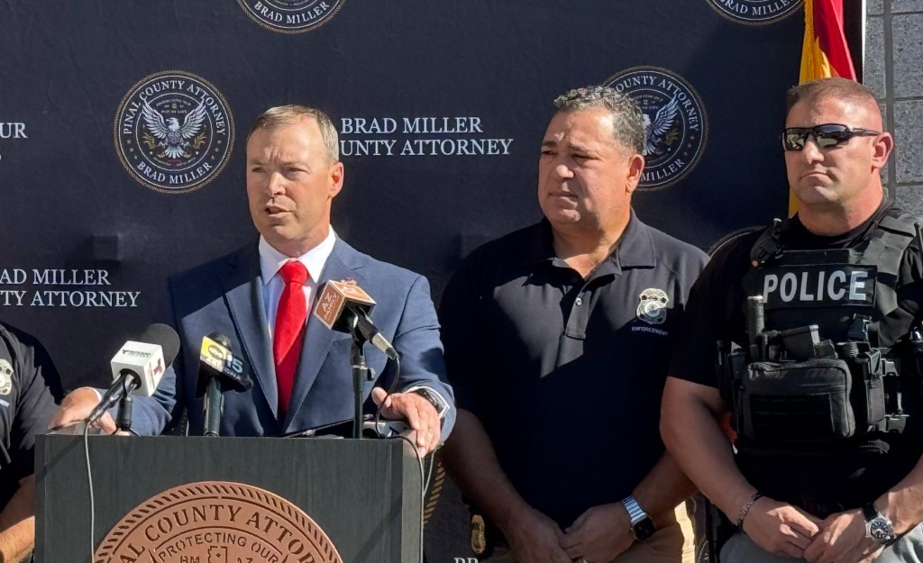 a man in a suit speaks at a podium, flanked by two other men -- one in a polo and another in a police vest