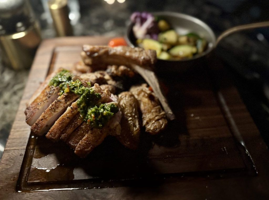 A tomahawk pork chop arranged, with crispy fingerling potatoes and a small skillet of steamed vegtables, on a cutting board.