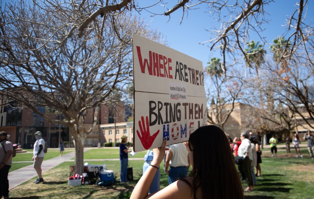 an anti-ICE protester holds a sign that says "where are they now? bring them home"