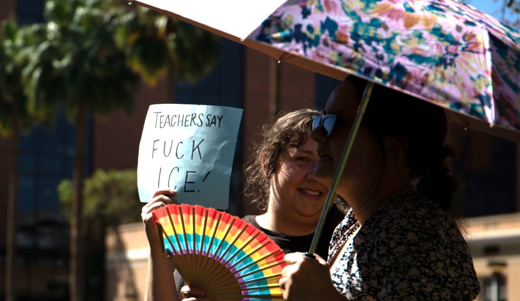 two women under an umbrella on a sunny day. one holds a rainbow paper fan and a sign that says "teachers say Fuck ICE"