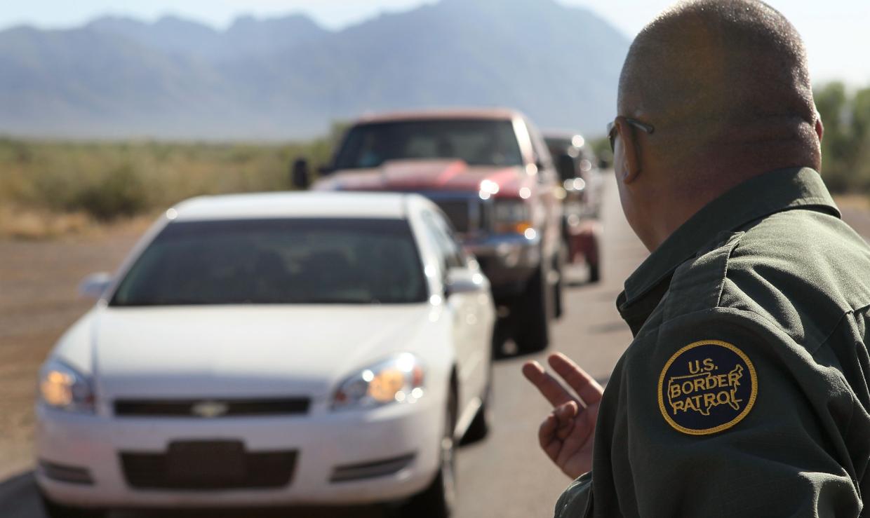 a border patrol agent waves forward a line of cars in a desert landscape