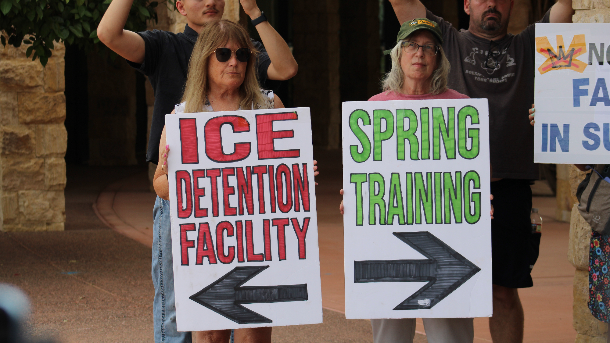 two proteters hold signs with arrows pointing in opposite directions. one says "ice detention center," while the other says "spring training"