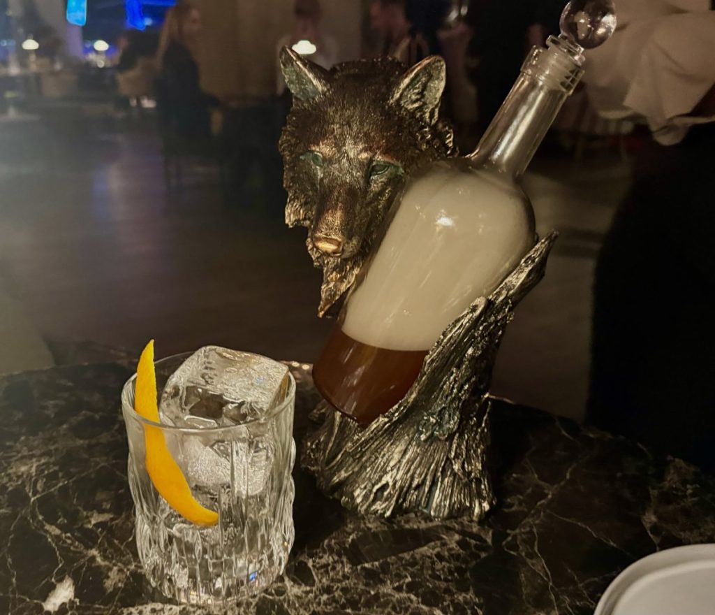 A cocktail glass next to a Wolf-shaped bottle holder containing a smoke-filled glass bottle.