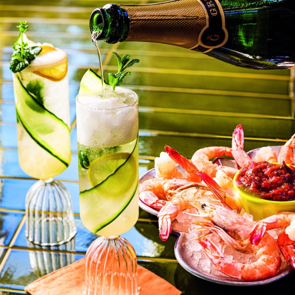 Sparkling wine is poured atop a cocktail sitting on a table next to a platter of shrimp cocktail.