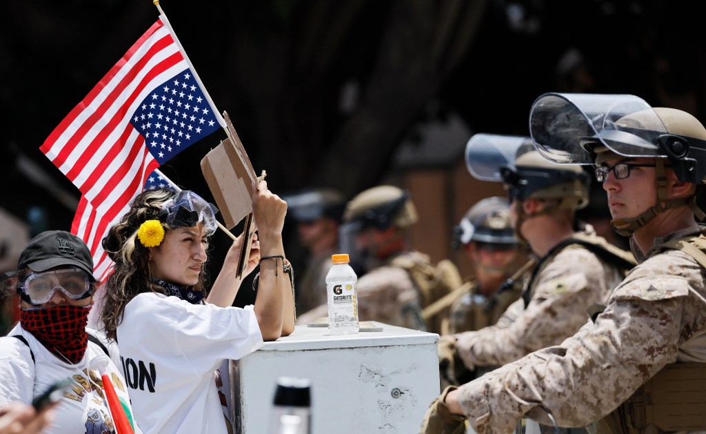 marines in tan fatigues stand in front of protesters waving upside down american flags