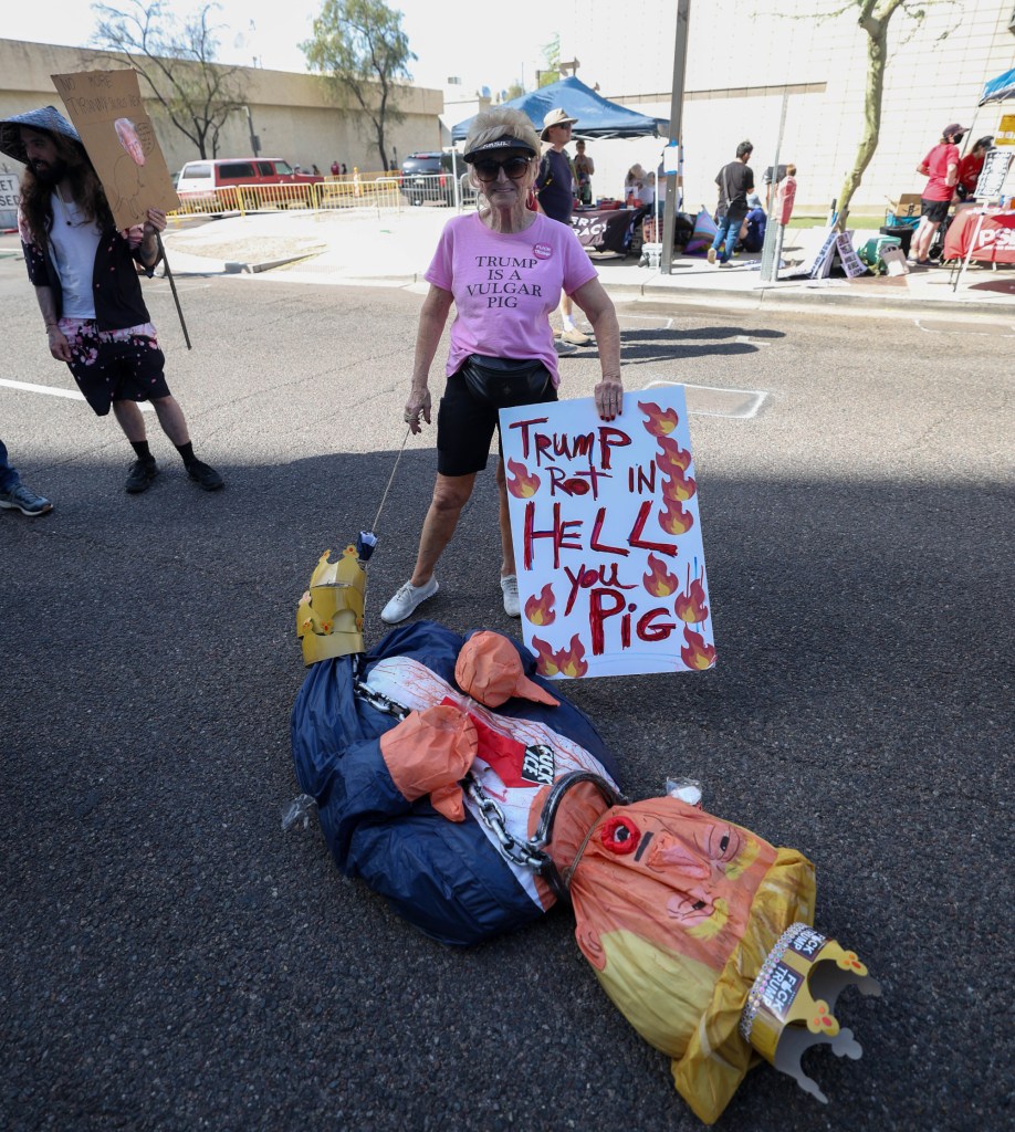 a protester stand over an effigy of donald trump on the ground while holding a sign that says "trump rot in hell you pig"