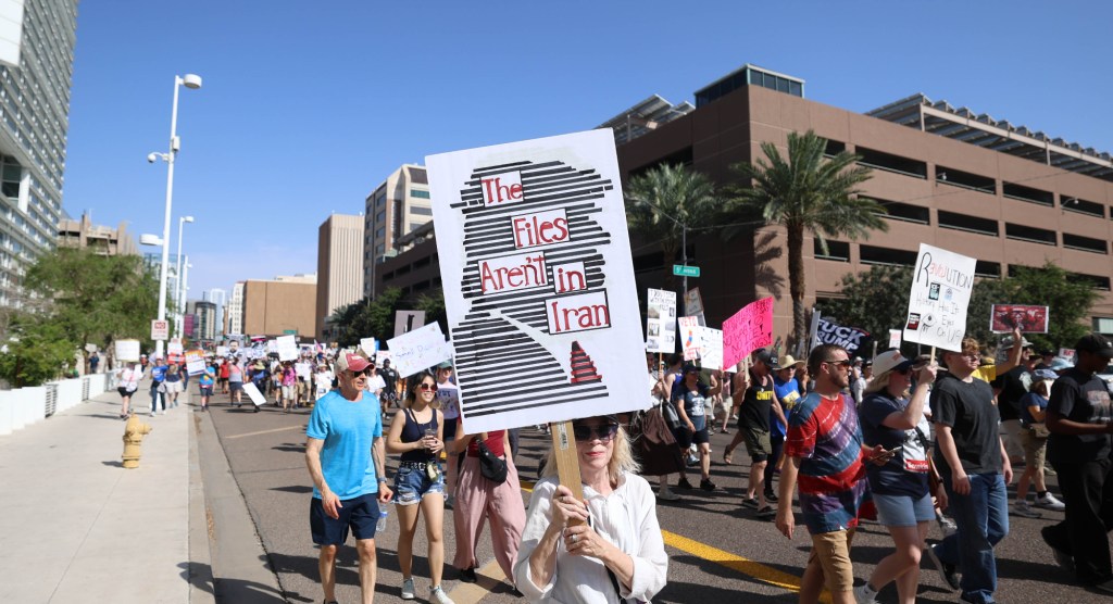 a protester holds a sign saying "the files aren't in iran" in a silhouette of donald trump made out of redaction lines