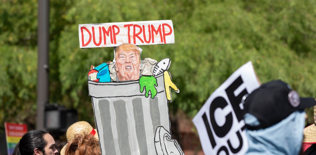 a protester holds up a sign with an illustration of donald trump in a metal trash can