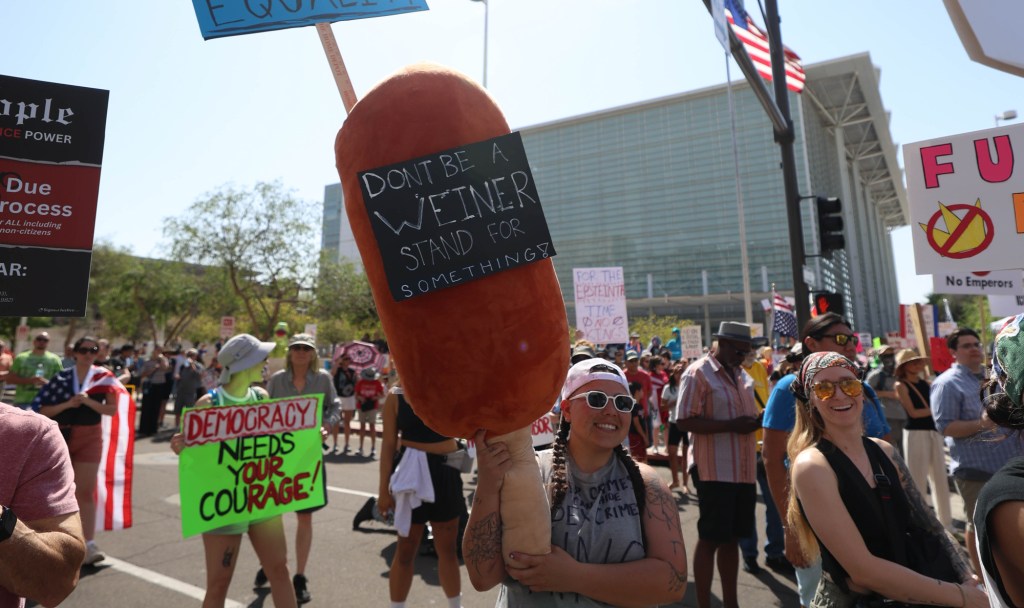 a protester holds a massive corndog, on which is written "'don't be a wiener, stand up for something"