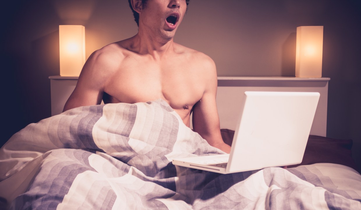 a shirtless man making an orgasmic face while sitting under his bed covers, with an open laptop on his lap