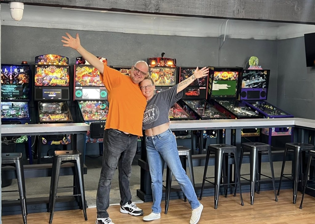 Jason and Tricia Webb stand inside Tilt Gamez in Glendale, surrounded by pinball machines at their arcade that opened in November 2025.