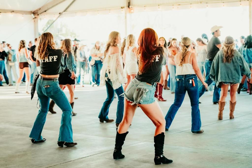 Two women in jean shorts and black t-shirts lead a line-dancing session in an outdoor tent at the Boots in the Park music festival.