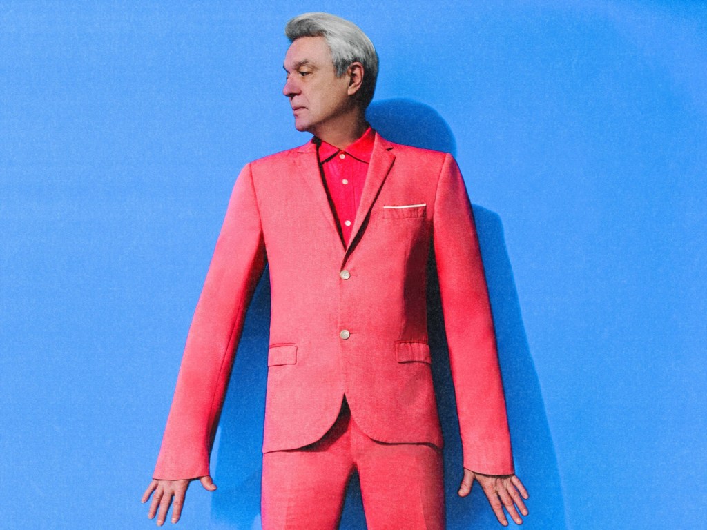 David Byrne in a pink suit standing against a blue background in a promotional photo for his 2026 tour.
