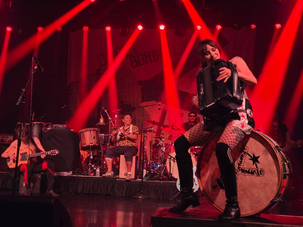 Erica Mancini plays accordion on a bass drum in front of two other musicians at a Gogol Bordello set