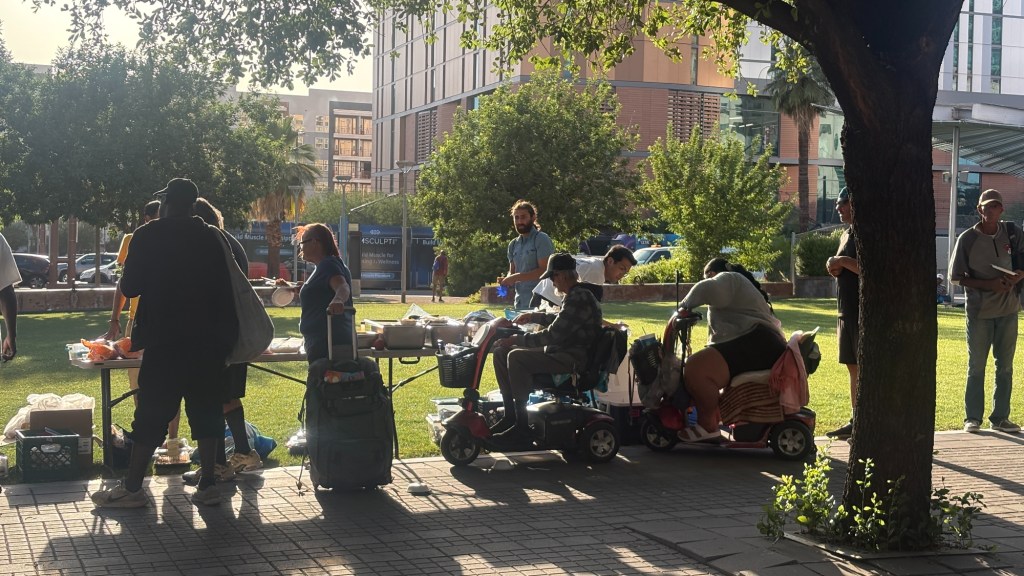people lined up outside on a sunny day by tables with food on them