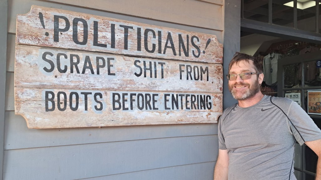 gary swing next to a sign that says "politicians! scrape shit from your boots before entering"