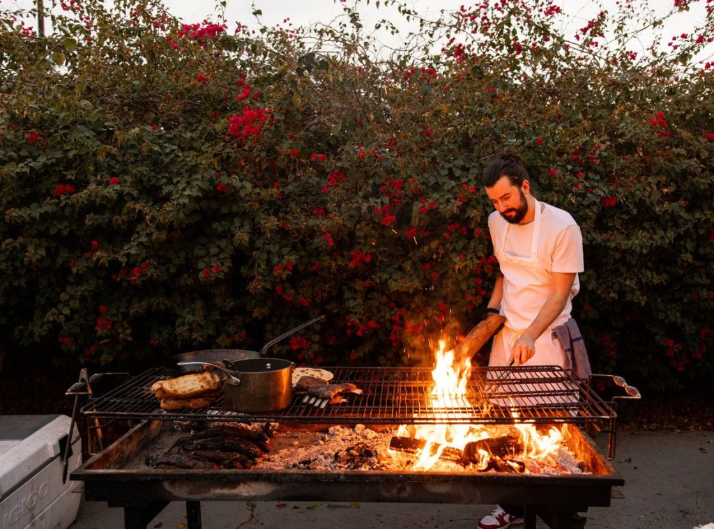 A chef tends the fire of a large grill outside.