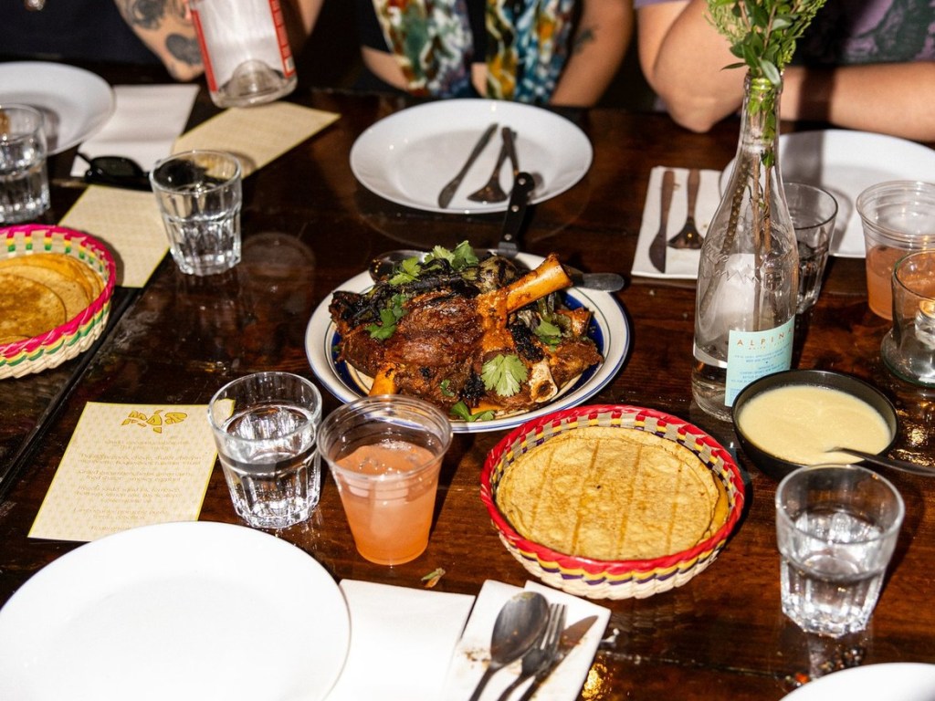 People sit around a table filled with a platter of meat and baskets holding tortillas.