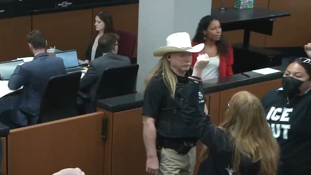 protesters in "ice out" shirts raise fists as they leave a arizona senate committee room. a long-haired man in a cowboy hat and police vest watches them