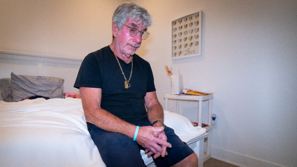 michael marion sits on the edge of a bed, wearing a balck shirt