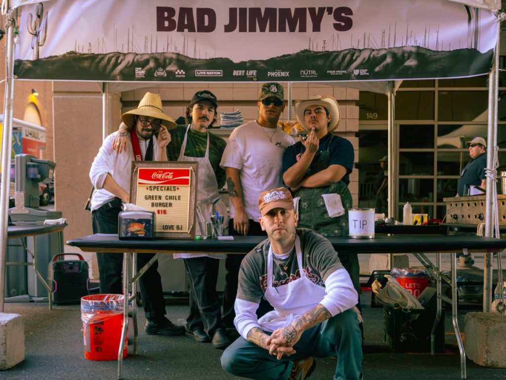 Bad Jimmy's owner James Piazza kneels in front of a VIVA PHX food booth with his team.