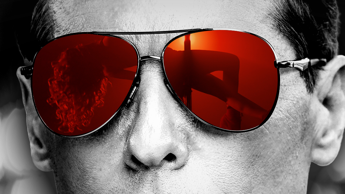 a black and white close up of a man's face. he wears sunglasses, which are in color and show the reflection of an exotic dancer on a stripper pole