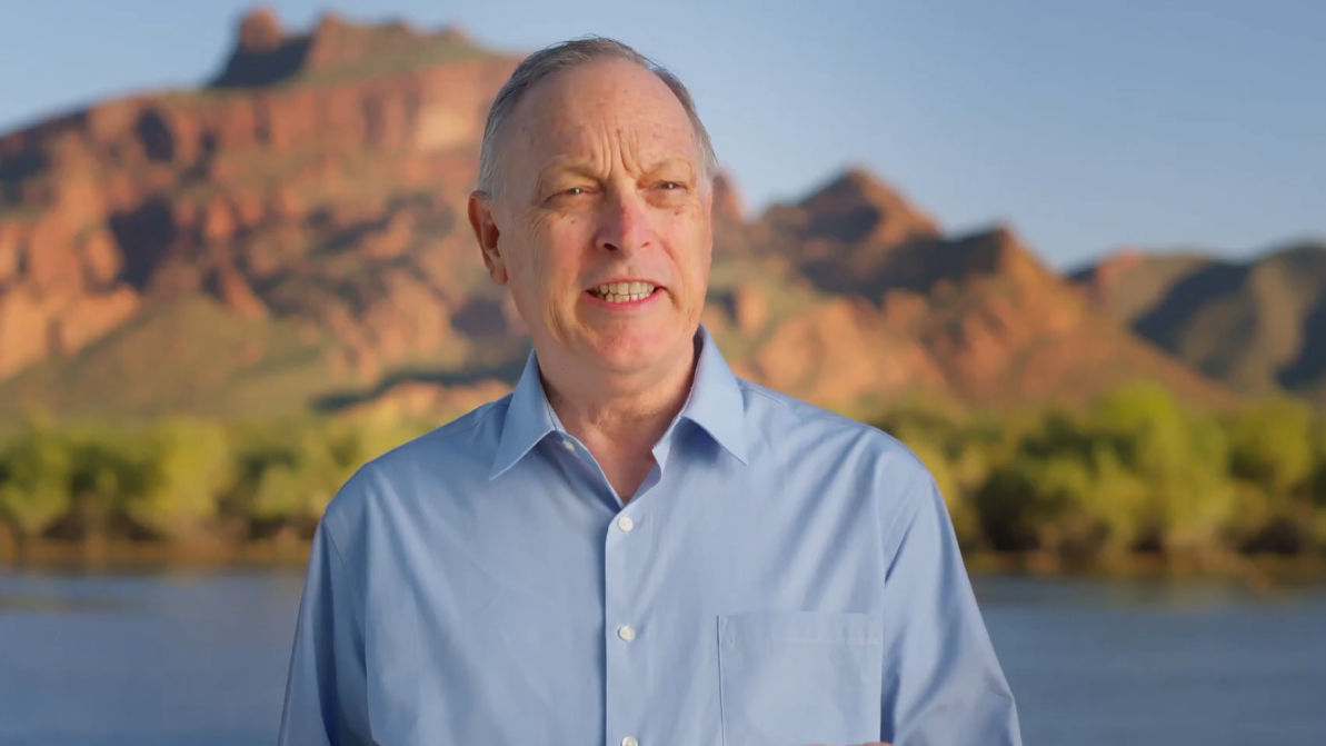 andy biggs stands in front of a mountain and a body of water