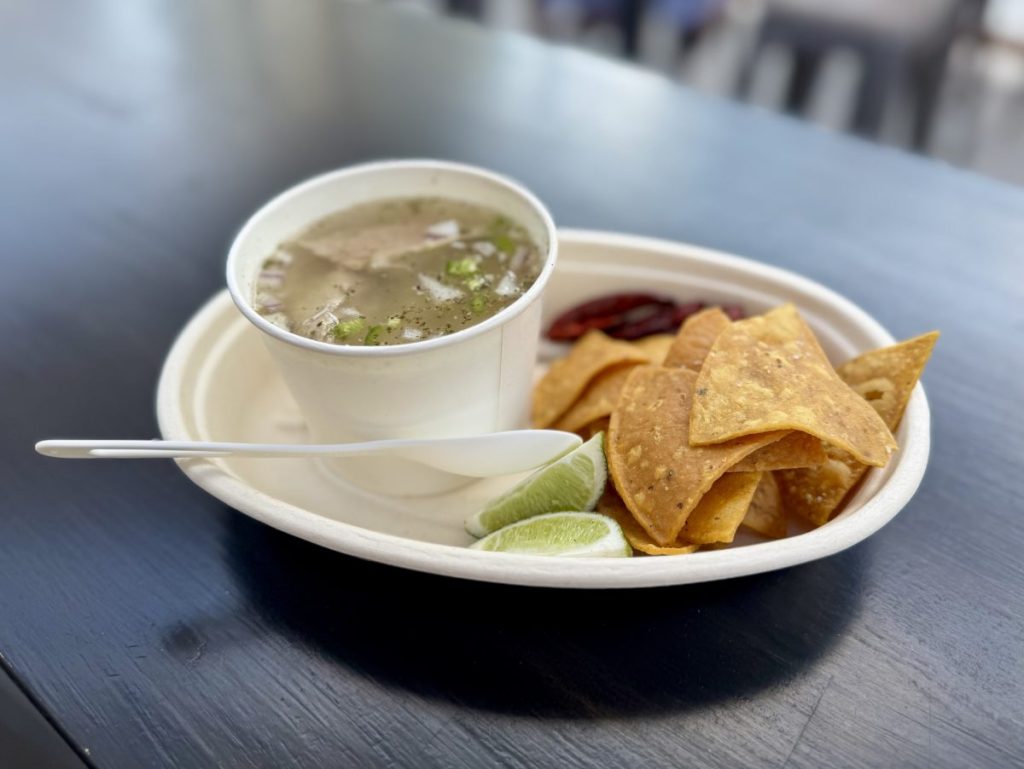 A cup of Pozole on a plate with chips, chiles and lime wedges.