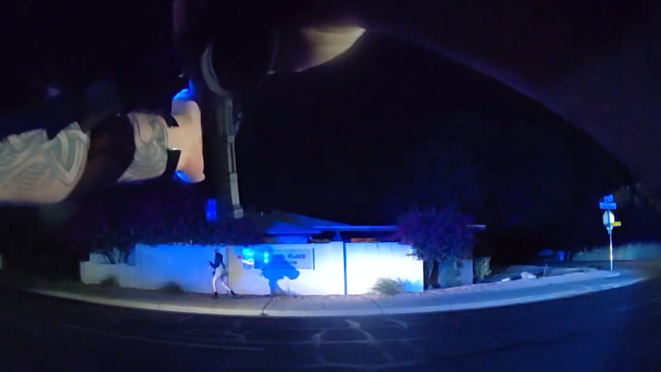 a body-cam still showing hands pointing a rifle at a person walking on a sidewalk at night
