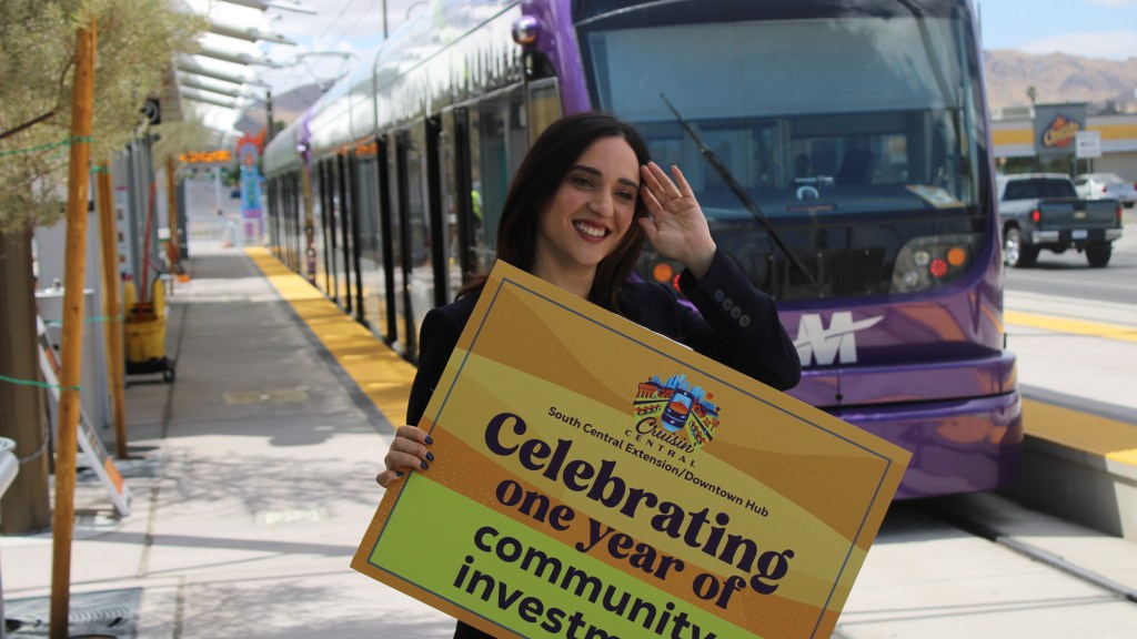 yassamin ansari holds a sign in front of a valley metro light rail train. the sign says "celebrating one year of community investment"