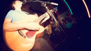 A musician and vocalist plays a guitar onstage at a music venue.