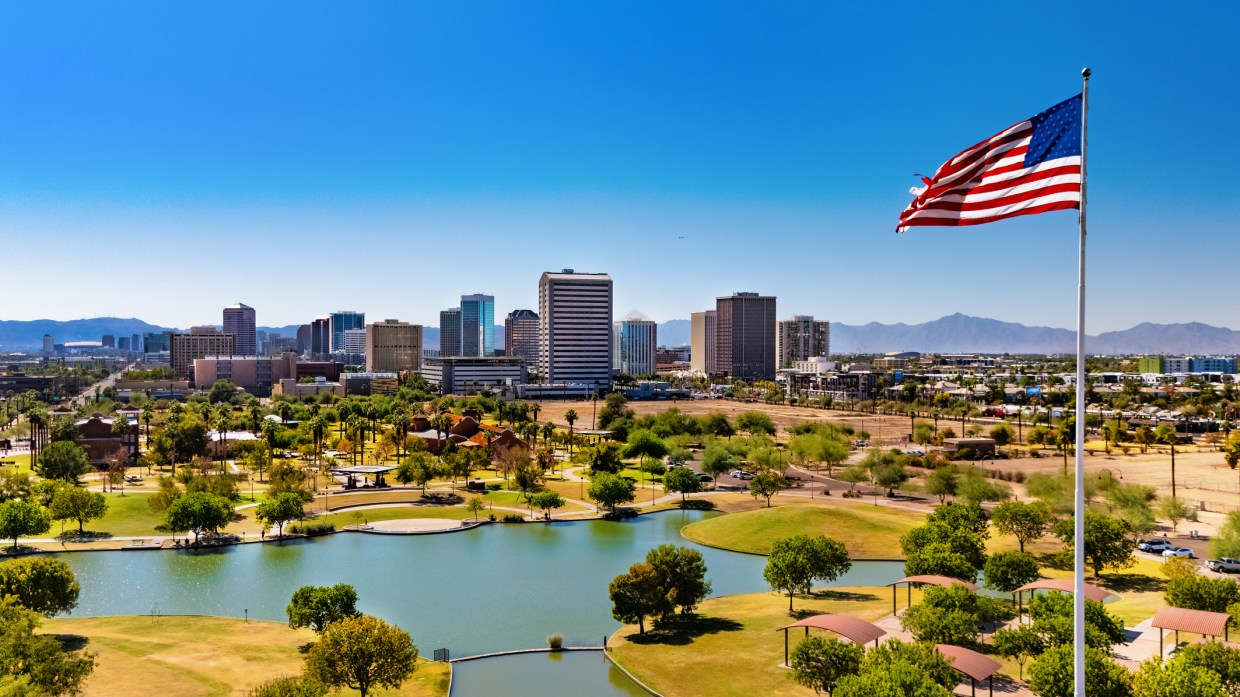 a view overlooking a pond at steele indian school park, beyond which are office buildings in midtown phoenix. an american flag flies on a pole.