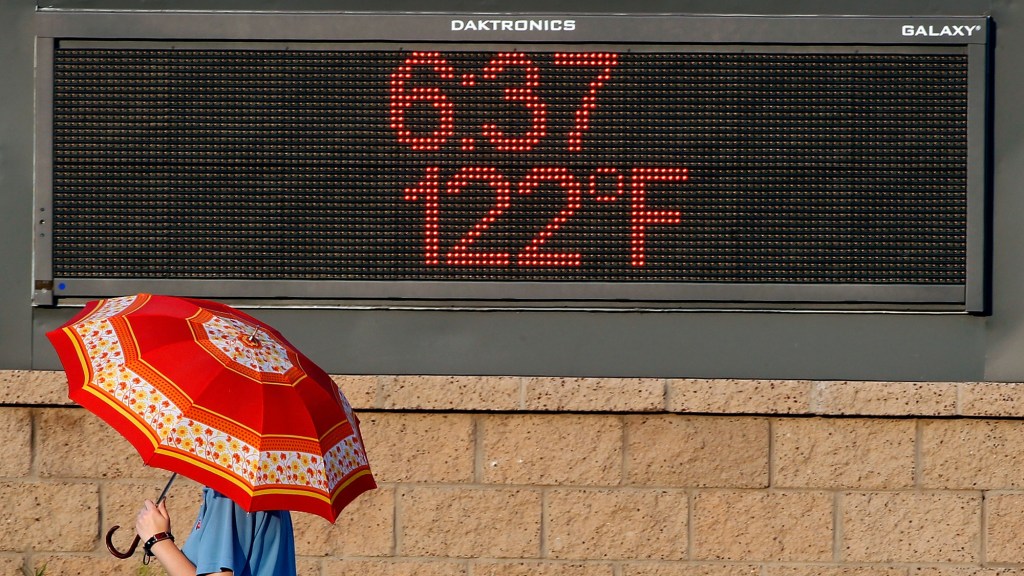 a person under an umbrella walks by a digital sign showing the temperature is 122 degrees at 6:37 pm