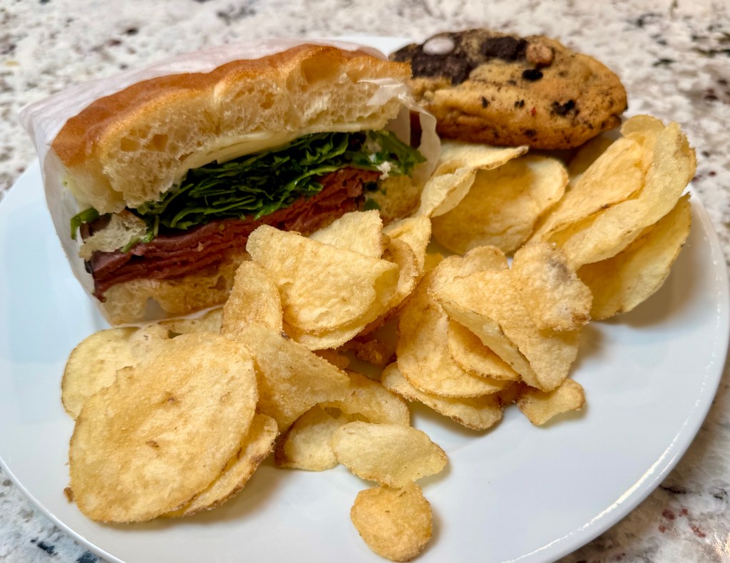 Half of a sandwich from Paper Bag Co. with chips and a Chewk's cookie on a plate.
