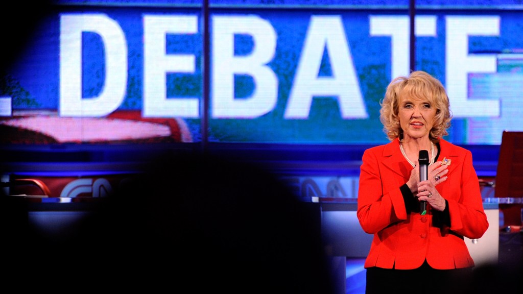 jan brewer stand with a microphone in a red blazer in front of a screen that says "DEBATE"